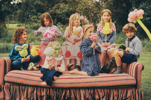 Six children sitting on a striped daybed in an outdoor garden setting, each holding a colorful balloon shaped like an animal, with a dog lying beside them.