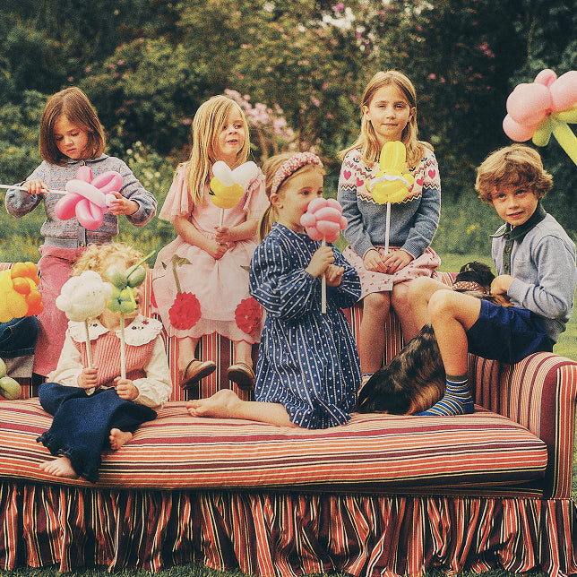 Five children sitting on a striped outdoor daybed holding pink, yellow, and white balloon animals, with a dog beside them and a garden background.