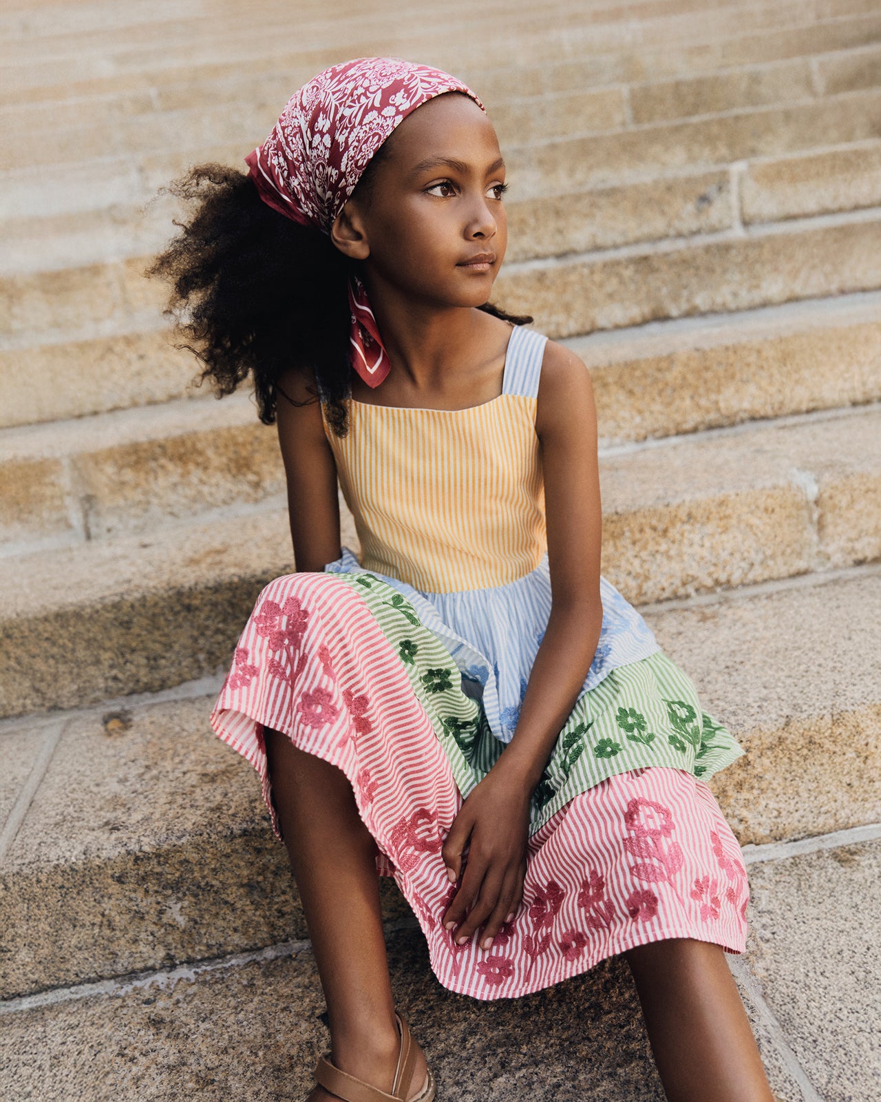 Young girl sitting on stone steps wearing a colorful dress with a headscarf.