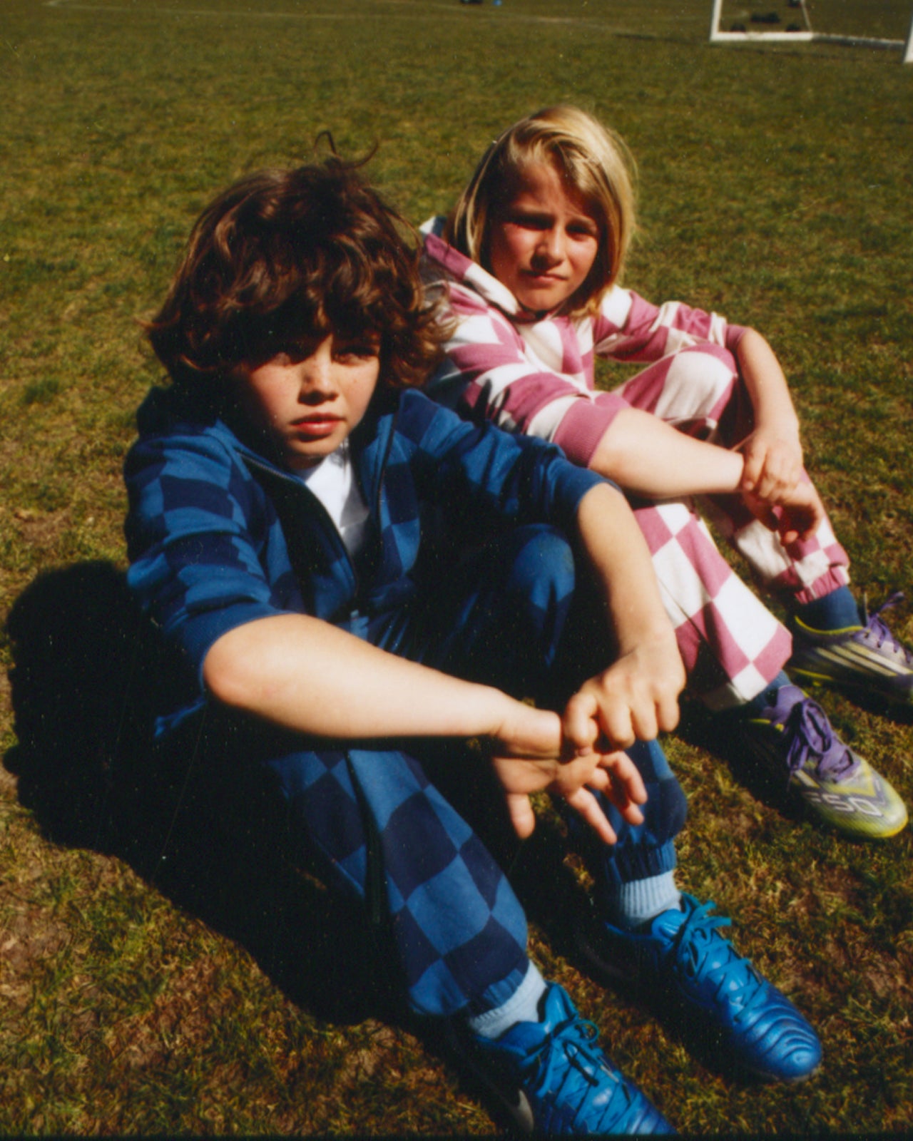 Two children sitting on grass, one in a blue checkered outfit and the other in a plaid outfit.