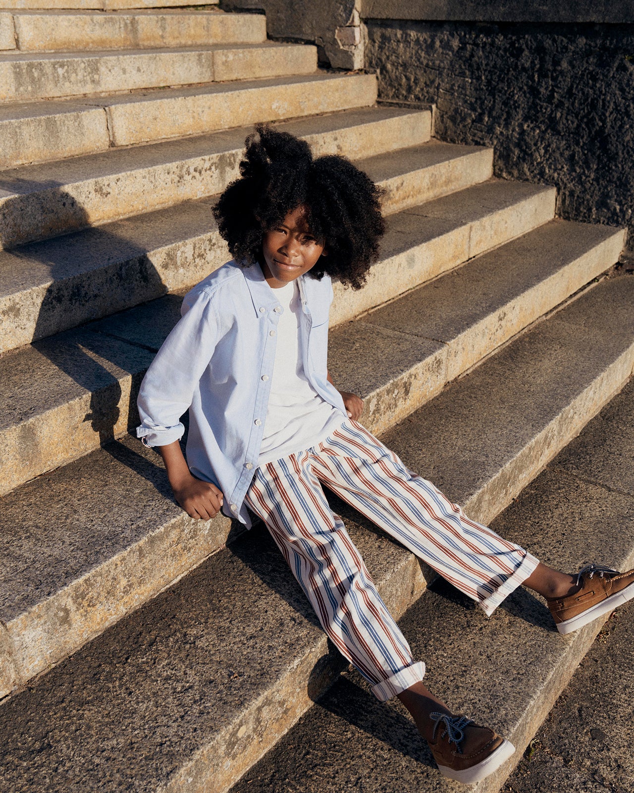 Child sitting on stone steps wearing a light blue shirt, white pants with stripes, and brown shoes.