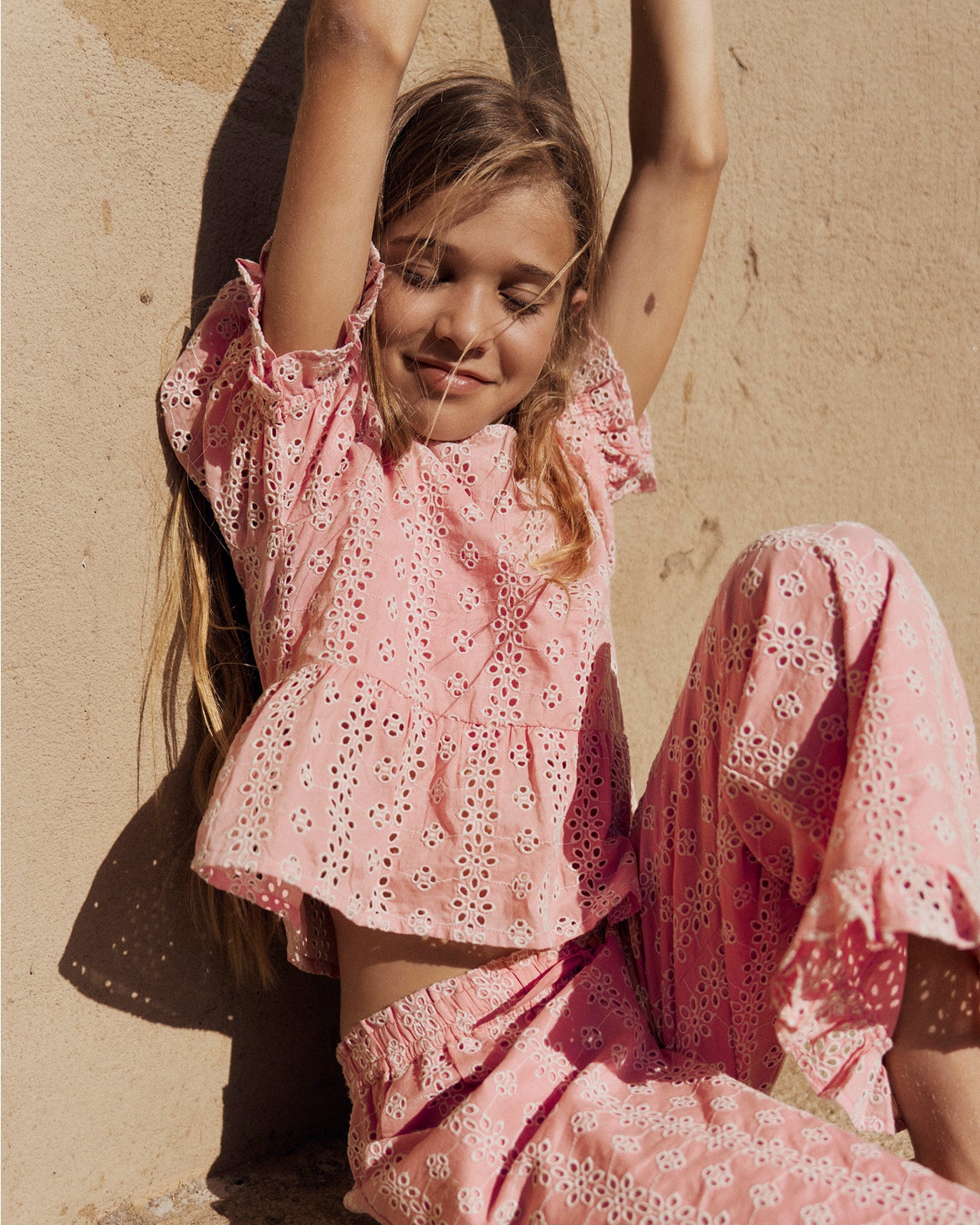 Young girl in a pink outfit sitting against a beige wall.