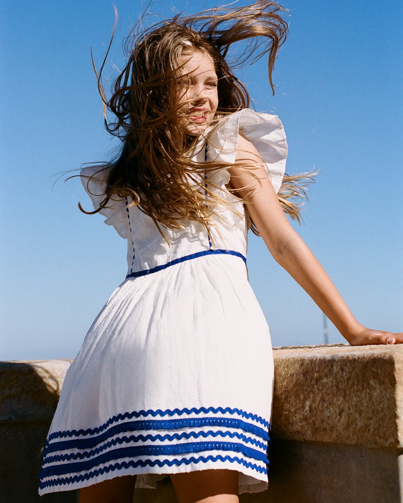 Young girl in a white dress with blue trim standing against a clear blue sky.