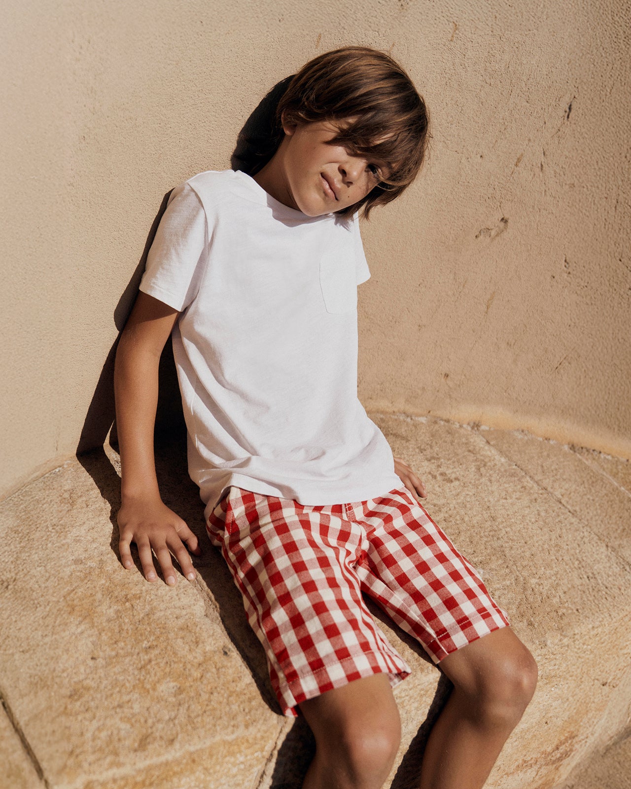 Child wearing a white t-shirt and red checkered shorts against a beige wall.