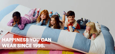 Children playing on a colorful bed with text 'Happiness you can wear since 1995'.