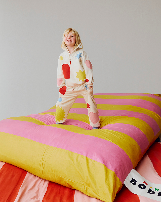 Person wearing a colorful onesie standing on a bed with pink and yellow striped bedding.