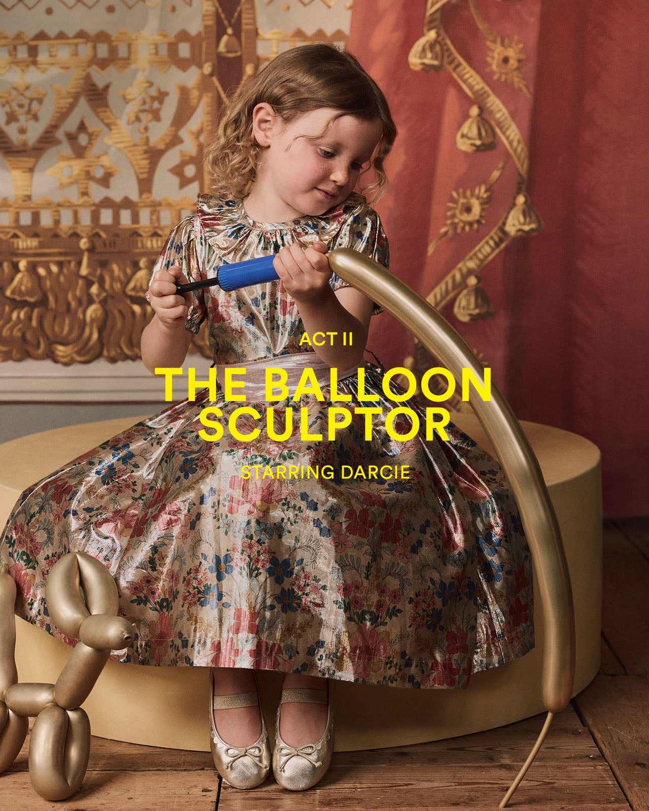 Young girl in a floral dress working with balloons against an ornate wall.