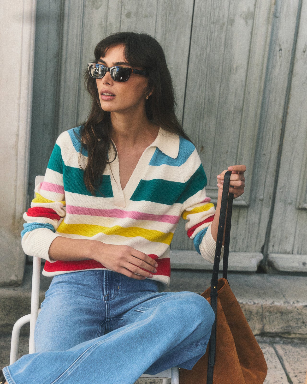 Woman wearing a colorful striped sweater and sunglasses, sitting on a chair.