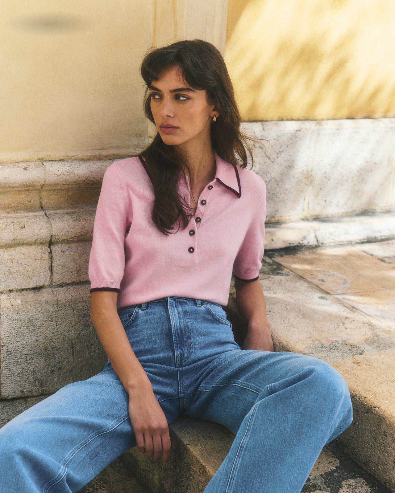 Woman wearing a pink polo shirt and blue jeans sitting on stone steps.