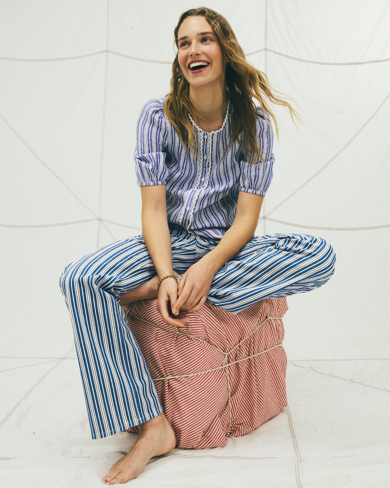 Woman sitting on a pink ottoman wearing a blue and white striped outfit in a tiled room.