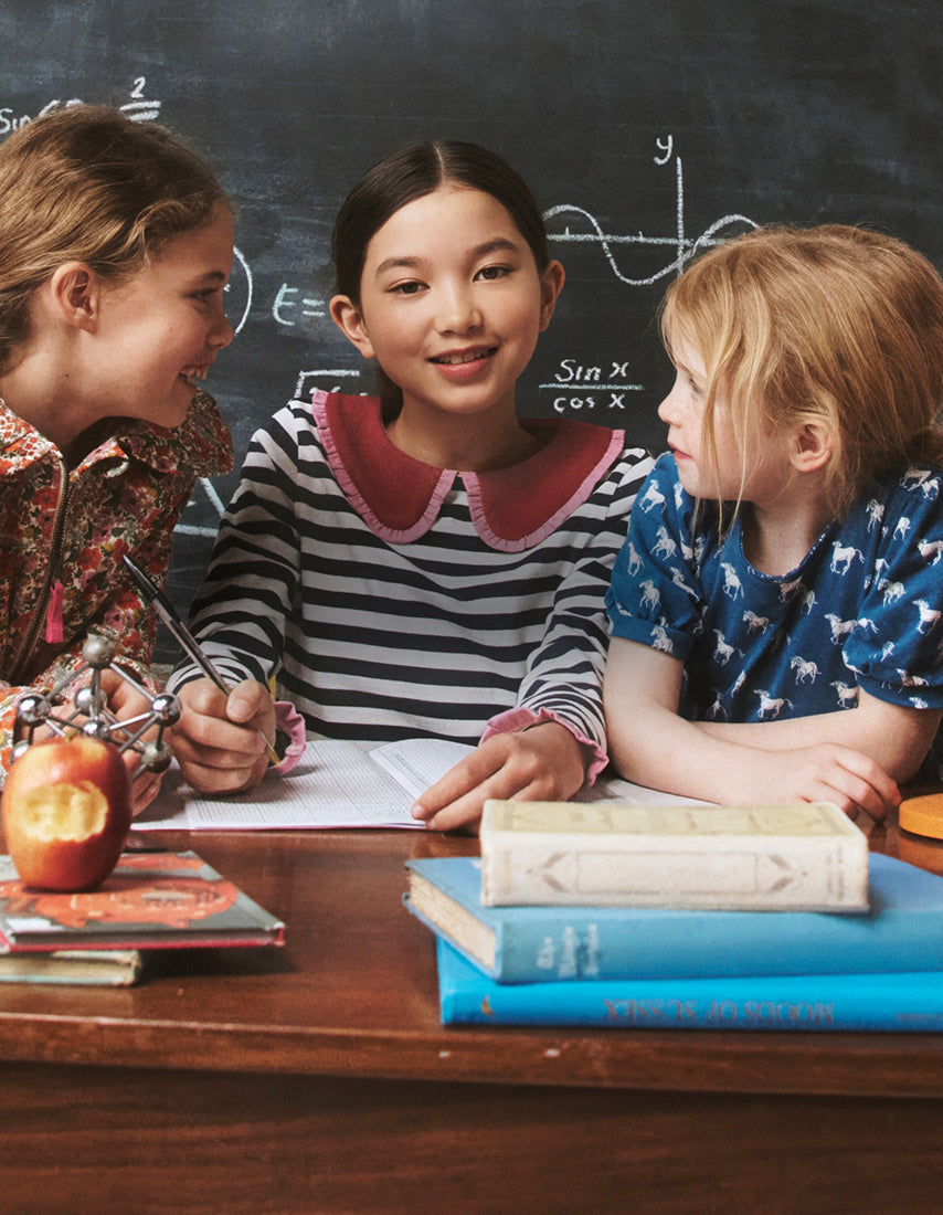 Three children sitting at a desk with books and an apple, in front of a chalkboard.