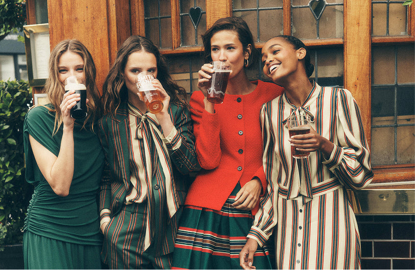 Four women standing together outdoors in front of a wooden building, each holding a pint of beer. They are wearing colorful, patterned outfits including a dark green dress, striped blouses, and a bright red cardigan.