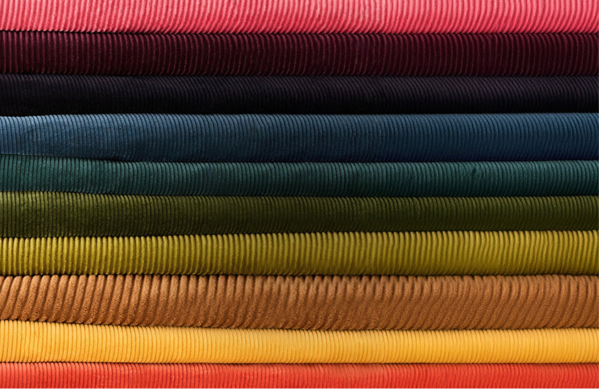 A close-up stack of ribbed fabric or material samples in multiple colors, including pink, navy, teal, green, olive, tan, mustard, yellow, orange, and red, arranged horizontally.