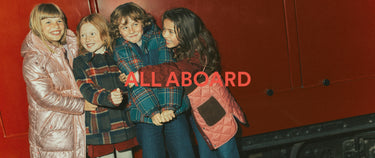Four children hugging in front of a red train, wearing colorful winter jackets. A red text overlay reads 'ALL ABOARD'.