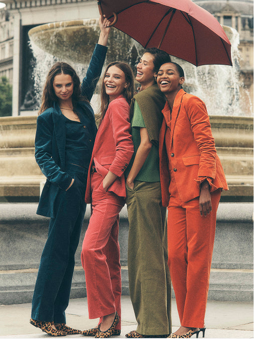 Four women posing together in front of a stone fountain, each wearing a different vibrant tailored pantsuit. The suits are in deep teal, coral pink, olive green, and bright orange.
