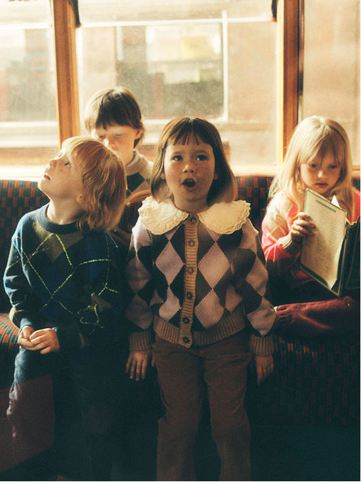 A vintage-style photograph showing four children sitting on a train or bus. The central child wears a brown and beige argyle sweater with a white lace collar, while the child on the left wears a dark blue argyle sweater, and the child on the right holds a book.