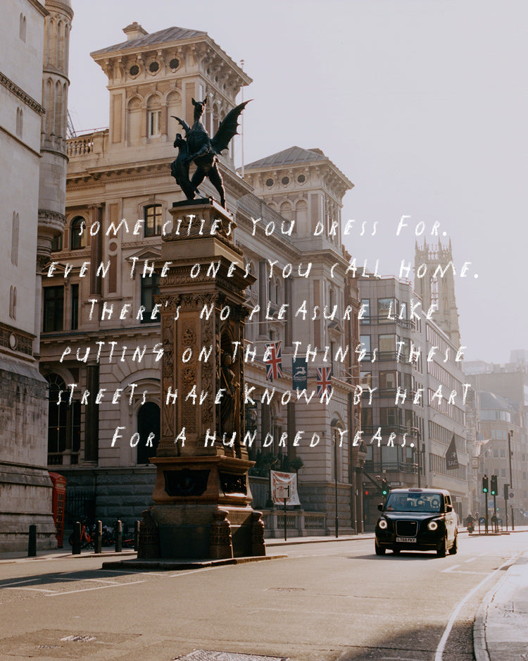 A scenic photograph of a London street with a central statue on a pedestal, historic buildings, a black cab, and overlaid white handwritten text reading, 'SOME CITIES YOU DRESS FOR, EVEN THE ONE'S YOU ALL HOME. THERE'S NO PLEASURE LIKE PUTTING ON THE THINGS THESE STREETS HAVE KNOWN BY HEART FOR A HUNDRED YEARS.