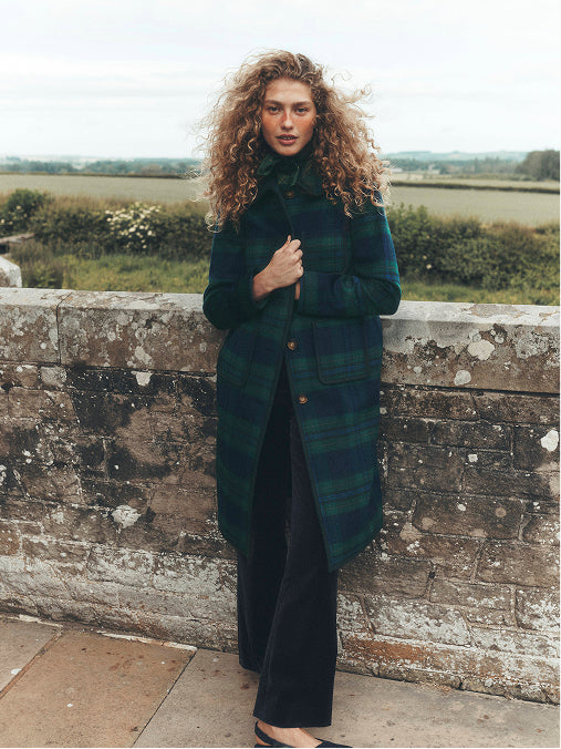 A woman with long curly hair stands outdoors against a weathered stone wall, wearing a dark green and black plaid overcoat over a black outfit.