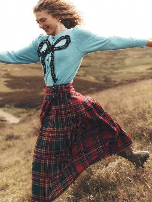 A woman outdoors in a grassy field wearing a light blue sweater with a black lace bow detail and a red tartan plaid skirt.