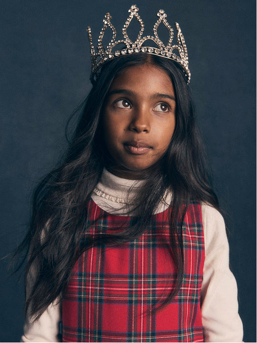 A young girl with long dark hair wearing a silver tiara with rhinestones, a red and navy plaid pinafore dress over a cream turtleneck, photographed against a dark slate blue background.