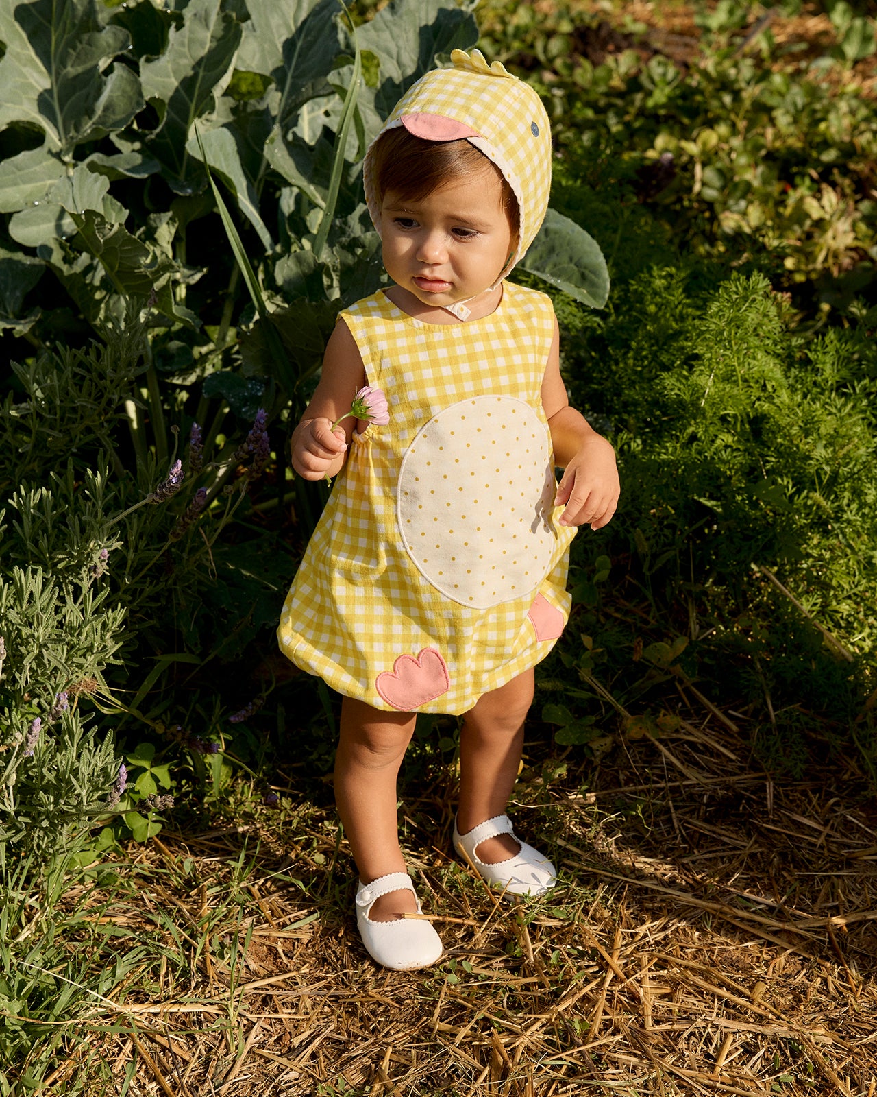 Child in a yellow checkered dress and bee hat standing in a garden