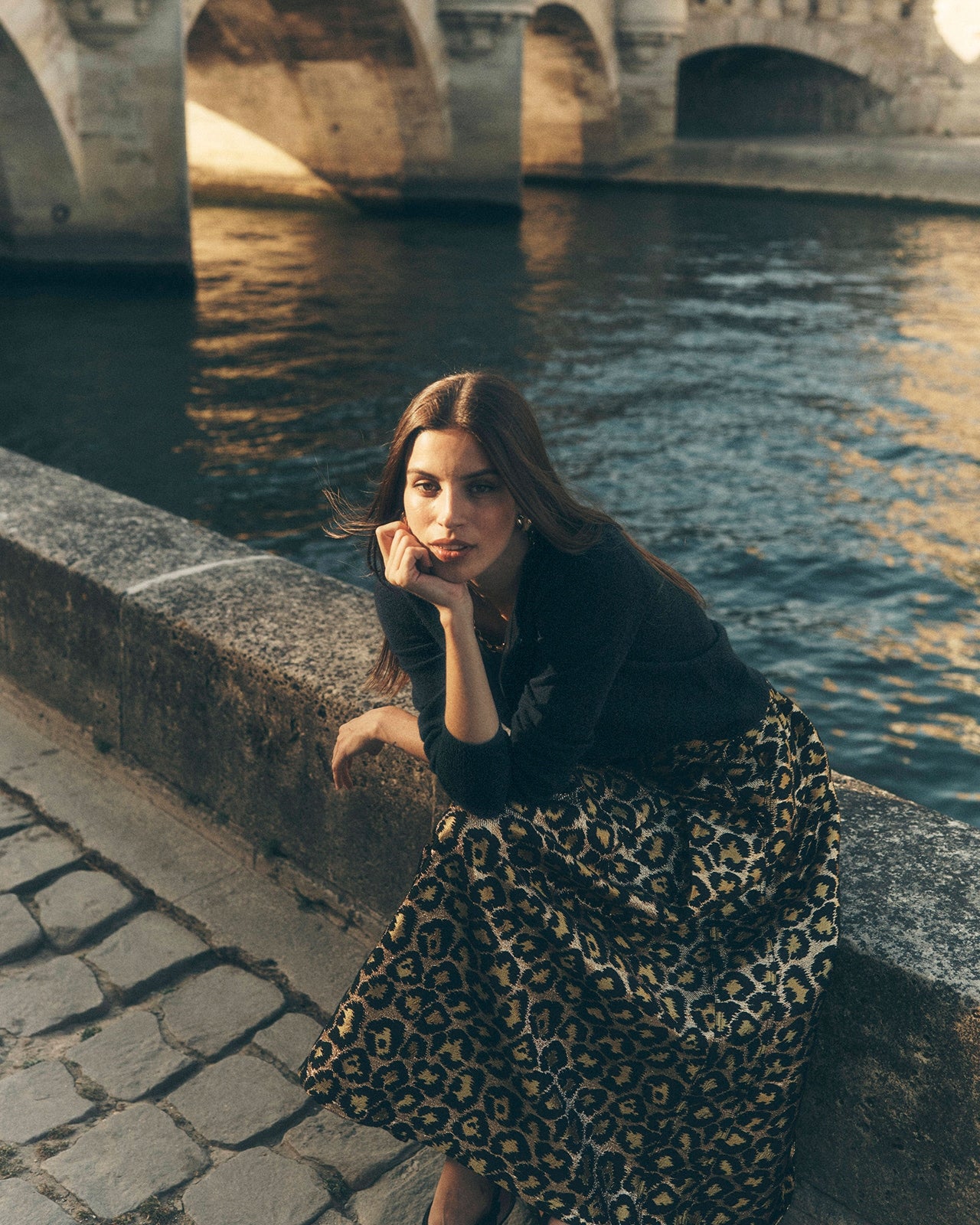 Woman sitting by a river with a stone bridge in the background